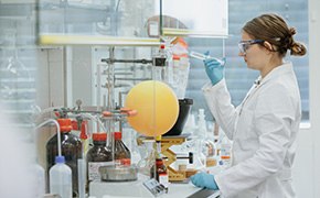 A scientist wearing a white lab coat, safety goggles, and blue gloves works in a laboratory using a pipette. In front of her is a round-bottom flask with an orange substance connected to glassware for a chemical experiment. Various bottles and lab equipment are visible on the workbench.