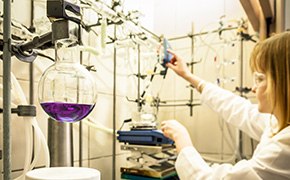 A laboratory setting where a scientist is conducting an experiment. A round-bottom flask containing purple liquid is prominently displayed, while the scientist is using a pipette to add a substance from a nearby apparatus. The background features a complex arrangement of glassware and tubing, indicating a detailed chemical process.