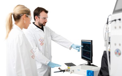 Process Analytical Technology (PAT tools) Two scientists in lab coats and gloves working with laboratory equipment. One is pointing at a computer screen displaying graphs, while the other looks on attentively. The scene takes place in a modern laboratory environment.