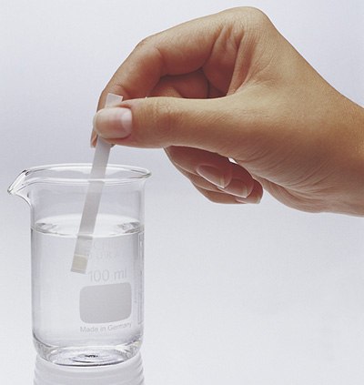 MQuant® test strip in beaker Mano sumergiendo una tira reactiva en un vaso de precipitados de vidrio transparente lleno de agua. El vaso está marcado con medidas que indican 100 ml y lleva la etiqueta "Made in Germany". La acción sugiere un proceso de pruebas químicas o de calidad.
