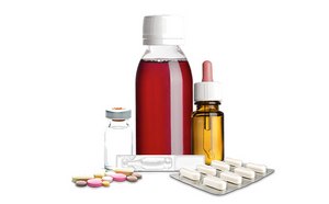 Surtido de artículos médicos: una botella de medicina líquida roja con una tapa blanca, un pequeño vial de vidrio con una tapa de metal, un cuentagotas con líquido de color ámbar, y varias píldoras y cápsulas, algunas sueltas y otras en blísteres.