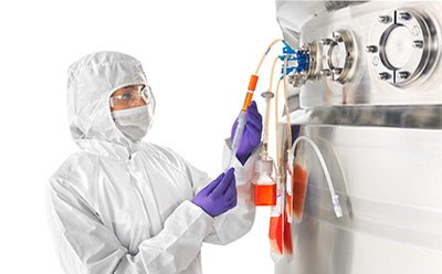 Individuo con equipo de protección blanco de cuerpo entero, incluidos la redecilla para el pelo y guantes morados, que está trabajando en una máquina industrial. La persona está manejando dos tubos de plástico flexible, uno naranja y otro azul, conectados a una máquina de acero inoxidable con un medidor circular y varios conectores.