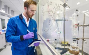 <p>Copper-Catalysed Carbon-Heteroatom Bond-Forming Processes</p>
 A laboratory technician in a blue lab coat and purple gloves is taking notes in front of a fume hood. Various glassware, including a round-bottom flask and a separating funnel, is visible on the counter, along with chemical structures and diagrams on a whiteboard in the background.