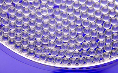 Primer plano de una placa de Petri circular llena de numerosas microesferas o gotitas esféricas de gel transparente, dispuestas muy juntas sobre una superficie lisa. La composición general enfatiza la uniformidad y la precisión de la disposición de las microesferas.