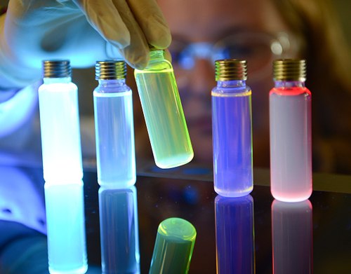 Vials of OLED materials lit up by UV light La imagen representa materiales emisores de luz para diodo orgánico emisor de luz (OLED). Una persona que lleva guantes sostiene un vial verde brillante entre otros viales brillantes de colores brillantes sobre una superficie reflectante. Los viales emiten varios colores: azul claro, verde amarillento, morado fuerte y rojo brillante. La cara de la persona y las gafas protectoras están sutilmente delineadas por el brillo, y cada vial tiene una tapa metálica. En la superficie puede verse el reflejo de los viales brillantes.