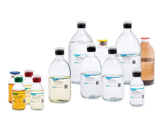 A collection of laboratory bottles containing various sterility testing culture media rinse and fluid. There are large transparent bottles with blue labels in the back row, medium-sized transparent and amber-colored bottles with different colored caps in the middle, and small transparent bottles with red, green, yellow, and blue caps in the front row. Each bottle has a label indicating its contents or purpose within sterility testing procedures.