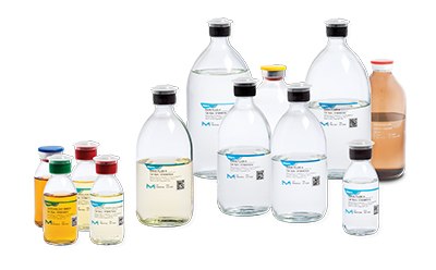 Conjunto de botellas de laboratorio que contienen varios medios de cultivo para ensayos de esterilidad, enjuagues y líquidos. En la fila de atrás hay botellas transparentes grandes con etiquetas azules, en el centro se observan botellas transparentes de color ámbar y tamaño mediano con tapas de diferentes colores, y en la fila delantera hay botellas transparentes de pequeño tamaño con tapas rojas, verdes, amarillas y azules. Cada botella tiene una etiqueta que indica su contenido o propósito dentro de los procedimientos de ensayos de esterilidad.