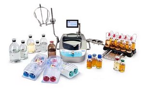 Surtido de fungibles, medios e instrumentos para ensayos de esterilidad. En el centro, hay un instrumento para ensayos de esterilidad con una pantalla digital y un tubo conectado. Alrededor del instrumento hay varias botellas de diferentes tamaños que contienen líquidos transparentes y de color ámbar. También hay varias placas de Petri con medios de colores en tonos azules, rojos y claros. A la derecha de la imagen, se observa una gradilla con viales de tapas rojas, que contienen posiblemente muestras o medios de cultivo. El fondo es blanco.