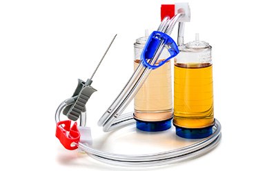 Sistema de filtración a través de membrana Steritest NEO. Este sistema consiste en una unidad de filtración con dos cámaras conectadas llenas de un líquido amarillento. La cámara izquierda tiene una abrazadera roja y la cámara de la derecha la tiene azul; ambas están montadas en soportes. También se observa una aguja unida a otro tubo.