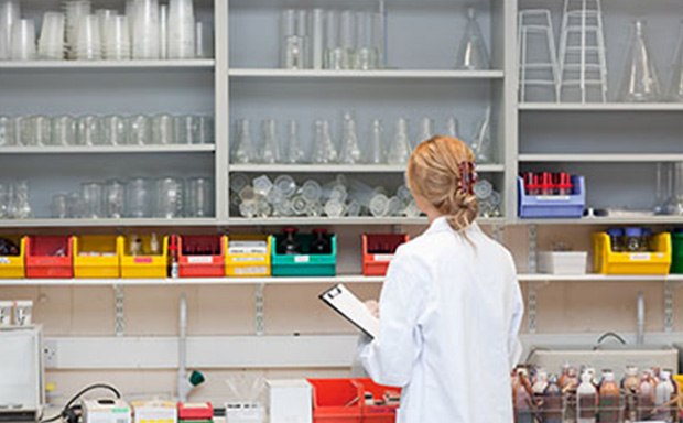 Lab worker looking at inventory on shelves A person in a white lab coat is standing in a laboratory facing away from the camera, holding a clipboard. The laboratory is equipped with shelves stocked with various lab equipment such as beakers, flasks, test tubes, and bottles containing chemicals of different colors. There are also racks of test tubes and other scientific apparatus on the bench in front of the person. The setting suggests a professional research environment, highlighting the meticulous organization and variety of tools used for scientific experiments.