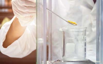 A person wearing a white lab glove is holding a spatula with a yellow substance over a glass beaker inside a laboratory fume hood. A person wearing a white lab glove is holding a spatula with a yellow substance over a glass beaker inside a laboratory fume hood.