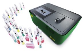 La imagen muestra un analizador verde y negro, posiblemente un espectrofotómetro, con una serie de tubos de ensayo coloridos delante, cada uno tapado y etiquetado con códigos de barras.