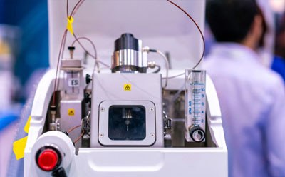 Mass Spectrometer Vista detallada de una máquina de espectrometría de masas de proteínas, con varios componentes como tubos, cables y diales, con un botón de parada de emergencia visible y una etiqueta amarilla de precaución. Al fondo, un científico con bata de laboratorio trabaja dentro de un entorno de laboratorio, destacando el uso de la máquina en la investigación científica y el avance tecnológico.