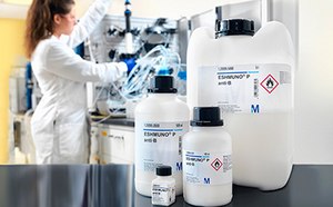 Científico con bata de laboratorio que trabaja con equipos y productos químicos en un entorno de laboratorio. En primer plano se observan tres recipientes químicos con etiquetas, dos son grandes y uno más pequeño, marcados con símbolos de peligro por sustancias inflamables y corrosivas.
