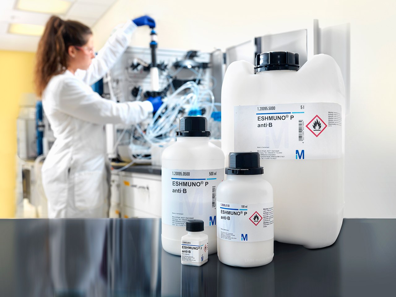 Affinity chromatography with Eshmuno® resins Entorno de laboratorio que muestra las herramientas y los productos químicos utilizados en la cromatografía de afinidad. En primer término, tres botellas etiquetadas con símbolos de peligro indican la presencia de sustancias inflamables y corrosivas, esenciales para interacciones bioquímicas selectivas y procesos de purificación. En el fondo, un científico en bata de laboratorio está ocupado en equipos complejos, probablemente relacionados con la fabricación a gran escala y el desarrollo de procesos en cromatografía de afinidad.