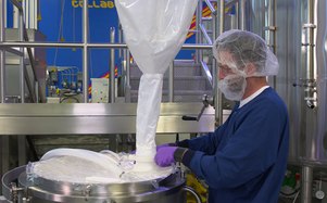En la imagen, una persona con ropa azul y una red para el cabello está vertiendo una sustancia blanca de una bolsa grande en un cuenco mezclador industrial en una instalación de fabricación.