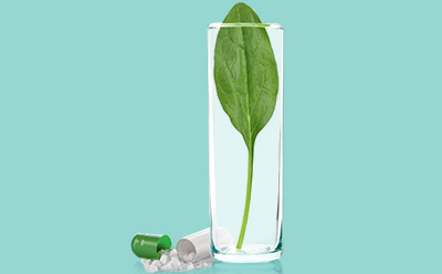 Folate | Mobius® fluid transport and storage systems La imagen representa un vaso transparente que contiene una gran hoja verde, rodeado de pastillas y cápsulas dispersas sobre un fondo verde azulado. Una de las cápsulas está abierta, revelando su contenido en polvo.