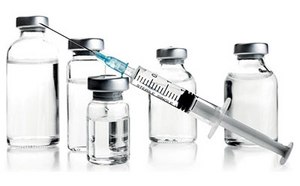 Una jeringa médica con aguja, parcialmente llena de líquido, está colocada en diagonal delante de cinco viales de varios tamaños con tapas metálicas sobre un fondo blanco.