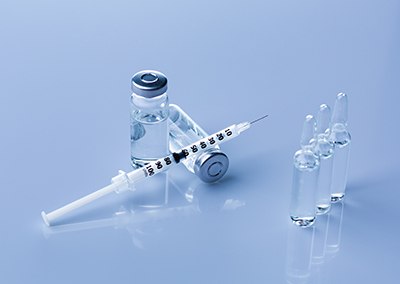 Parenteral drug formulation Entorno estéril en el que se muestran preparaciones farmacéuticas para inyectables y formulaciones parenterales. Pueden observarse los siguientes elementos: 1) Un vial transparente con un tapón precintado, que contiene una disolución líquida. 2) Al lado del vial, una jeringa blanca con marcas de medición. 3) Tres ampollas transparentes de pie en la superficie reflectante. Todo el conjunto está colocado sobre una superficie reflectante.
