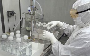 Técnico con un traje de sala limpia manejando medios para cultivo líquido de células en un laboratorio de bioprocesamiento, con filas de botellas cerradas cuidadosamente dispuestas que contienen los medios y el equipo esencial de laboratorio, al fondo, lo que indica un entorno estéril y controlado para aplicaciones de bioprocesamiento.