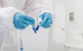 Persona con equipo de protección, que incluye guantes, manipulando una jeringa médica y un vial. El individuo está vestido de blanco, con guantes azules, y está concentrado en el equipo médico.