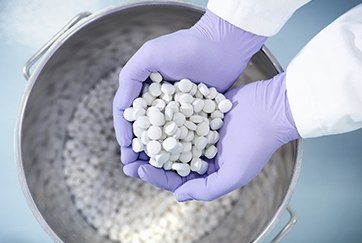 Solid formulation pills Un par de manos con guantes morados sostiene un puñado de comprimidos blancos y redondos sobre un gran recipiente metálico lleno de más pastillas. El fondo está suavemente difuminado, resaltando las manos y los comprimidos.