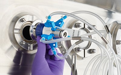 Una persona está conectando con cuidado un conector de varios tubos azules y blancos a un puerto metálico en una máquina. El individuo lleva guantes morados, lo que destaca la necesidad de precisión y cuidado en el manejo del equipo sensible. Los tubos blancos flexibles se prolongan fuera del marco, sugiriendo su conexión a un sistema más grande.