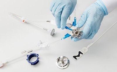 Manos cubiertas con guantes azules montando o desmontando meticulosamente un instrumento médico o científico. Los componentes (tubos, conectores y piezas especializadas de plástico y metal) están esparcidas sobre una superficie blanca. Una mano sostiene un componente con varias salidas, posiblemente para conexiones de tubo o cable. También se observa entre los artículos un objeto similar a una jeringa con una punta naranja.