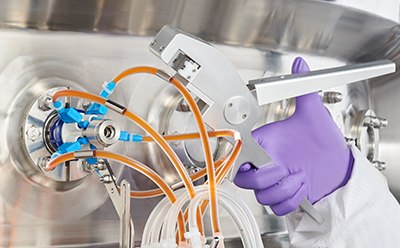 Persona con bata de laboratorio y guantes morados que parece estar ajustando el equipo con varios tubos conectados a él. El equipo consta de varios tubos naranjas y azules, lo que indica  o un aparato de análisis o transferencia de líquidos.