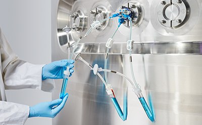 Persona con bata de laboratorio y guantes azules que maneja tubos transparentes llenos de un líquido azul brillante conectados a una máquina circular metálica con varios botones. La persona está llevando a cabo un experimento o proceso que implica la transferencia del líquido azul.