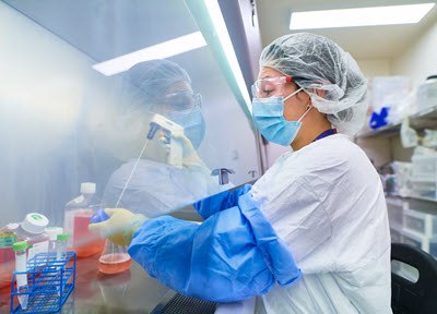 Viral Vector Upstream Platforms Available for Both AAV and LV Production operator in lab coat, mask and hood working in biosafety cabinet transferring cell culture media into a flask with pipette
