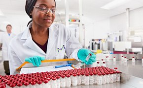 Bioactive Small Molecule Agonists Scientist looking at a vial in her hand. There is a collection of vials in front of her. In the background another scientist walks towards her.