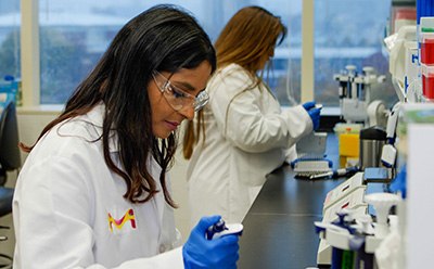 Bioactive Small Molecule Toxins Scientists at Bench A scientist sits at the lab bench wearing lab goggles, a lab coat, and gloves. She is pipetting. Behind her another scientist wearing a lab coat and goggles is using a multichannel pipettor. There are instruments and pipette tips on the right of the image.