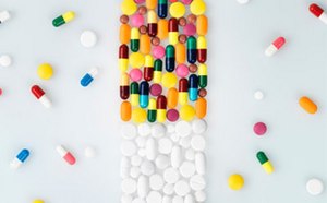 La imagen representa una variedad de comprimidos y cápsulas de diferentes colores dispuestos artísticamente sobre una superficie blanca imitando la forma de una botella de comprimidos, con el cuerpo compuesto de cápsulas multicolores y la tapa de comprimidos redondos blancos, todo rodeado de algunas pastillas sueltas.