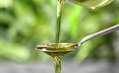 Olive Oil Pouring into Spoon La imagen parece mostrar un líquido viscoso y dorado, probablemente aceite, que se está vertiendo en una cuchara. El fondo verde borroso sugiere un entorno natural, que podría relacionarse con temas de cocina, ingredientes alimenticios, suplementos para la salud o productos naturales.