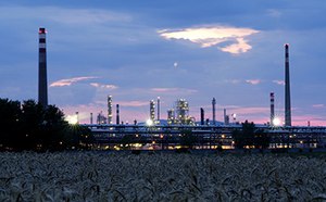 La imagen representa una planta industrial en el crepúsculo, con altas chimeneas y estructuras iluminadas contra un cielo nublado, vista sobre un campo de cultivos.