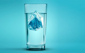 La imagen representa un vaso alto y transparente lleno de agua, con un gran cubito de hielo teñido de azul flotando en el centro sobre un fondo turquesa, que sugiere un ambiente fresco y refrescante.