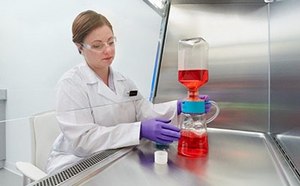 La imagen muestra a una mujer con bata blanca de laboratorio trabajando con un matraz de laboratorio que contiene un líquido rojo y está conectado a otro equipo con líquido azul en el fondo.