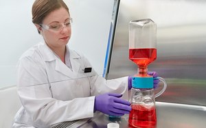 The image show a woman in a white lab coat, working with laboratory flask containing a red liquid, which is connected to another piece of equipment with blue liquid at the bottom.