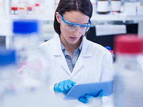 Scientist in a white lab coat and blue gloves uses a tablet in a laboratory, with reagent bottles blurred in the foreground.