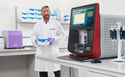 顕微鏡、CellASIC<sup>®</sup> ONIX2ライブセルイメージングシステム、コンピューターモニターが置いてあるベンチの前に座って微笑む科学者