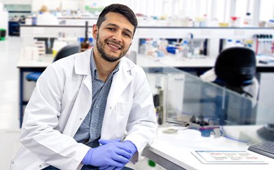 Scientist in a lab environment wearing a lab coat and protective gloves sitting at desk 
