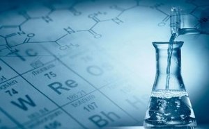 A laboratory scene featuring a clear glass flask filled with liquid, with a dropper positioned above it. In the background, a periodic table of elements is faintly visible, creating a blue-toned atmosphere that suggests a scientific research environment.