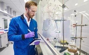 A scientist in a blue lab coat and purple gloves stands in a laboratory, holding a notepad and pen. They are positioned in front of a glass wall displaying laboratory equipment, including a round-bottom flask and a separating funnel. The background features various lab instruments and a clean, organized workspace.