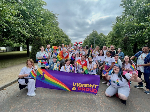 A group of about 30 employees holds up a banner that reads “Vibrant and Proud” with a rainbow flag on the side. They hold rainbow flags and many are smiling.