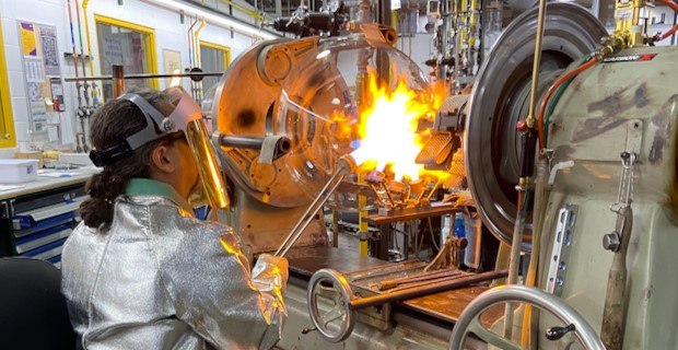 A round-bottom flask sits in a lathe and a glassblower directs a targeted flame onto the flask’s neck. The glassblower wears protective clothing, including a specialized jacket, gloves and face shield. 