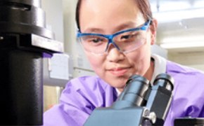 Female lab technician with Asian assemblance in lab protection cloth and working with a microscope.
