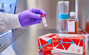 A laboratory scene showing a gloved hand holding a small vial with a red cap, next to a box with a geometric design and other laboratory containers in the background.