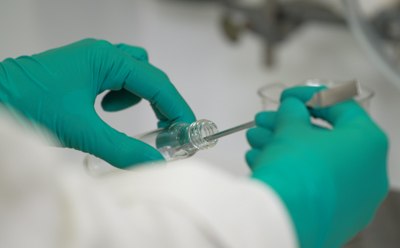 Pre-Clinical Lipids CDMO Services Two hands of an operator, clad in protective gloves, skillfully dispensing a fine white powder from a glass vial using a spatula.