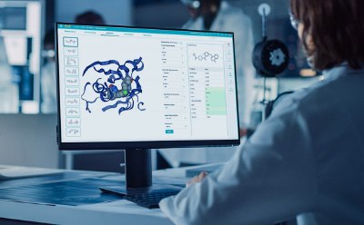 Benefits for Medicinal Chemists Científico en bata de laboratorio sentado ante un escritorio, interactuando con una pantalla de ordenador donde se observa una estructura molecular. En la pantalla se observa una representación tridimensional de una proteína o complejo, con varias herramientas y opciones de datos visibles en el lateral.